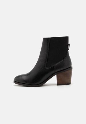 Bottine en cuir noir avec des panneaux latéraux élastiques, bout rond et un talon en bois texturé. Elle est dotée d'une languette à l'arrière.