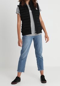 Black padded vest with hood over a gray T-shirt, paired with cropped light blue jeans and black sneakers, featuring frayed hems.