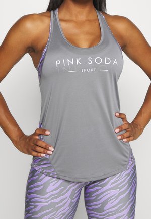 Vrouw die een grijs tanktop draagt met de tekst "PINK SODA SPORT", gecombineerd met paarse en grijs zebra-gestreepte leggings, handen in de zij.