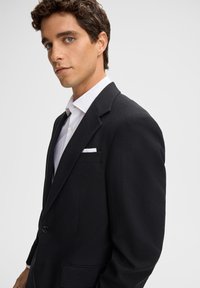 Blazer nero in lana con rever a punta, chiusura a un bottone e due tasche frontali. Indossato sopra una camicia bianca con bottoni.