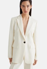 Blazer bianco realizzato in tessuto liscio, con rever a scollatura, singolo bottone nero e due tasche frontali. Vestibilità su misura con maniche lunghe.