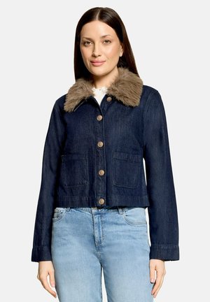 Frau trägt eine dunkelblaue Jeansjacke mit braunem Pelzkragen und goldenen Knöpfen, kombiniert mit hellblauen Jeans, stehend vor einem schlichten Hintergrund.