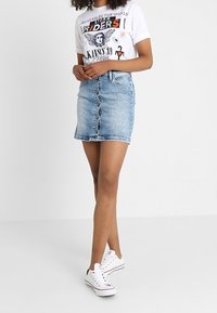 Bílý grafický tričko s multikolorovým potiskem, v kombinaci s světlou džínovou minisukní s předními knoflíky a roztrhaným lemem, nošeno s bílými teniskami.