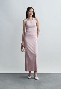 Γυναίκα με ροζ μακριά φόρεμα χωρίς μανίκια με ριμπ ύφασμα, κρατώντας μια μικρή μπεζ τσάντα και φορώντας λευκά παπούτσια με μυτερή μύτη, σε γκρι πάτωμα.