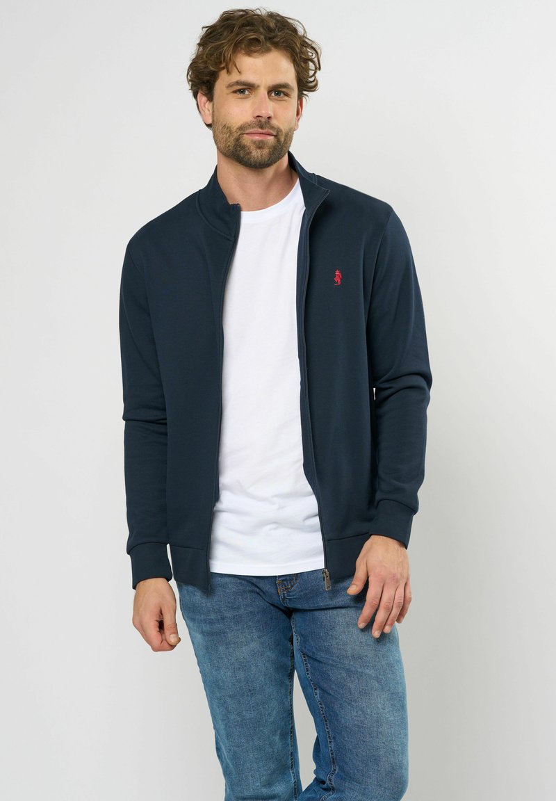 Marine blauwe zip-up jas met hoge kraag, voorzien van een rood logo op de borst, gecombineerd met een wit T-shirt en blauwe jeans. Glad textuur van de stof.