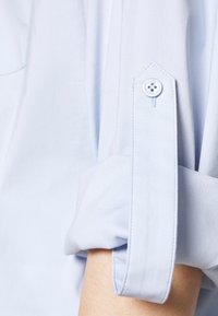 Light blue shirt sleeve rolled up and secured with a buttoned strap, showing part of the forearm.