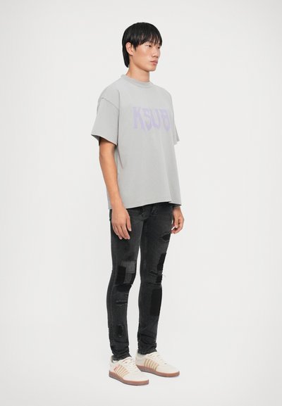 Grå oversized t-skjorte med "KSUBI" trykt i lilla, kombinert med svarte slitte skinny jeans og hvite joggesko med brune detaljer.