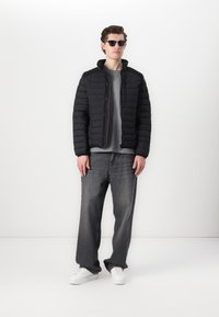 Black padded jacket with a high collar, worn over a gray shirt. Paired with wide gray jeans and white sneakers. Sleek sunglasses complete the look.