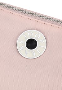 Borsetta di tessuto rosa con superficie testurizzata. Presenta un logo metallico circolare con incisione e un'apertura centrale nera, chiusa da una zip.