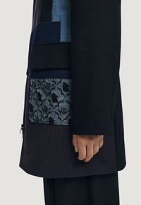Cappotto nero su misura con tessuti vari. Presenta una tasca blu scuro con motivo floreale, materiale testurizzato e dettagli a righe.