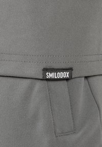 Grau Sportbekleidungsstoff mit glatter Textur, versehen mit einem sichtbaren Etikett mit der Aufschrift "SMILODOX" am Saum und dezenten Nähten.