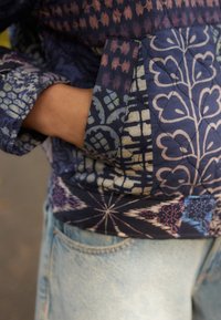 Persoon in een blauwe gestreepte jas met hand in zak en lichtblauwe spijkerbroek, met focus op het midden en de kledingdetails.