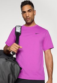 Lila rövid ujjú sportpóló, amelyen bal oldalon fekete Nike logó található, egy kéz tartja egy szürke táskaszíjat.
