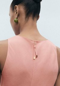 Abito senza maniche di colore rosa con una cerniera centrale sul retro, completato da un tirante in metallo dorato. Orecchini circolari verdi in vista.