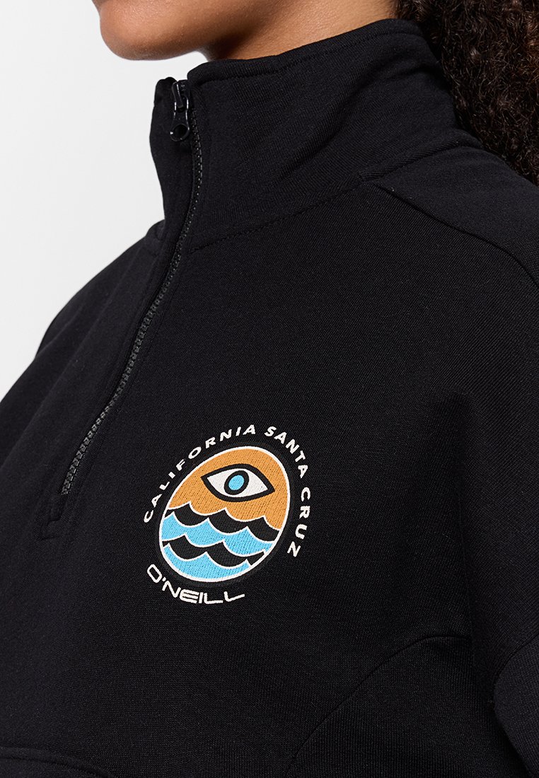 Sudadera negra de cuello alto con media cremallera, que presenta un logotipo redondo multicolor bordado con diseño de olas y un ojo en el pecho.