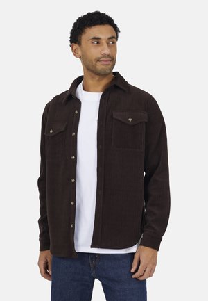 Man draagt een donkerbruine corduroy blouse met knopen over een wit T-shirt en blauwe spijkerbroek, kijkt iets naar de zijkant tegen een effen achtergrond.