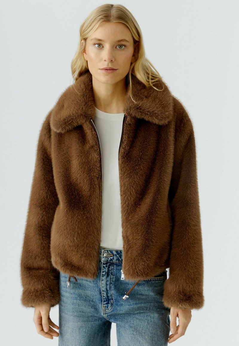 Vrouw met blond haar draagt een bruin, harig zip-up jack over een witte top en blauwe spijkerbroek, staand tegen een effen lichte achtergrond.