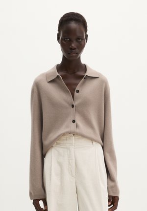 Model draagt een beige gebreid vest met knopen en kraag en crèmekleurige broek met hoge taille en wijde pijpen tegen een effen achtergrond.