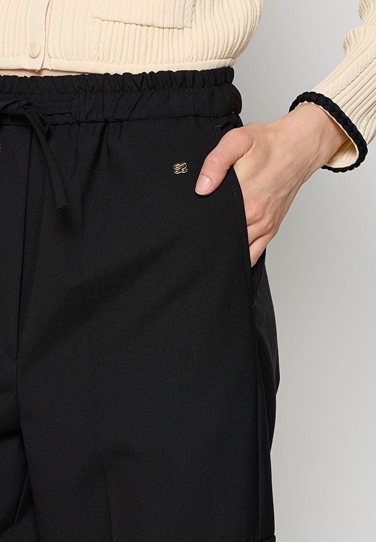 Zwarte broek met een elastische tailleband, voorzien van een trekkoord en zijzakken. Een klein gouden logo is zichtbaar aan de voorkant.