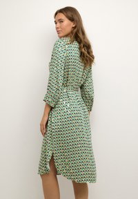 Rochie verde cu un model triunghiular repetat în nuanțe de albastru și verde închis. Prezintă talie cu brâu și mâneci până la trei sferturi. Detaliu cu fante pe laterale.