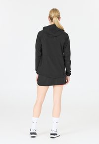 Svart idrottsjacka med huva, med ett strukturerat mönster och långa ärmar, kombinerad med en svart kjol och vita sportskor.