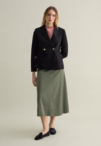 Blazer negro de doble botonadura con botones dorados, combinado con una blusa a rayas rojas y blancas, una falda verde de longitud media y unos zapatos negros sin cordones.