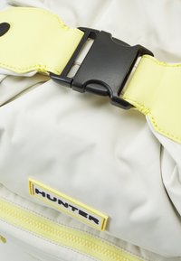 En närbild av en ljusfärgad väska med ett svart spänne och en gul rem, med en gul varumärkesetikett som läser "HUNTER."
