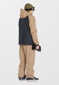 Beige en zwarte skijas met capuchon, gecombineerd met bijpassende baggy broek. De outfit heeft cargozakken en een gestructureerde gebreide olijfgroene beanie.