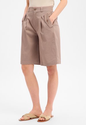 Kobieta nosząca beżowe, wysokie szorty do kolan z kieszeniami na patki zapiętymi na guziki, ręce w kieszeniach oraz beżowe, płaskie sandały z supełkiem.