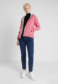 Roze lichtgewicht jack met een grijze capuchon, vooraan met ritssluiting en elastische manchetten, gecombineerd met donkerblauwe jeans en witte sneakers.