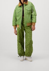 Grön bombarjacka med en brun imitation av pälskrage, dragkedjestängning och två bröstfickor, i kombination med gröna mönstrade cargobyxor och vita sneakers.