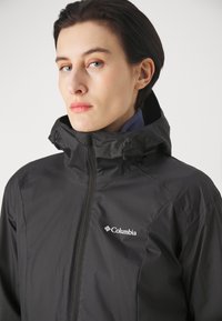 Black waterproof jacket with a high collar, full zipper, and adjustable drawstrings. Features a small white Columbia logo on the chest.