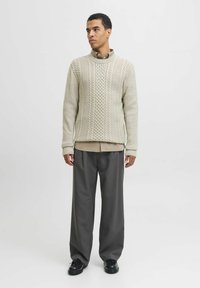 Pull en maille torsadée beige clair avec un motif texturé, superposé sur une chemise beige, associé à un pantalon large gris et des chaussures noires.