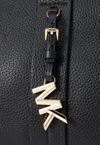Borsa a tracolla in pelle nera con una targhetta in tono oro che presenta iniziali stilizzate, cintura regolabile e accessori decorativi.