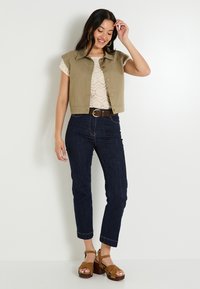 Chaleco sin mangas color caqui sobre una blusa de encaje color crema, combinado con jeans de mezclilla azul oscuro y sandalias de plataforma marrones.