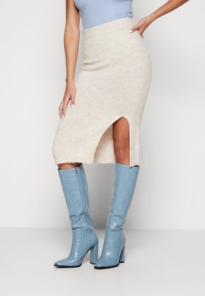 Vrouw draagt een beige gebreide midirok met een split aan de voorkant en lichtblauwe kniehoge laarzen met hoge hakken, poseert met één hand op haar heup.