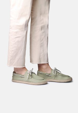 Pieds portant des chaussures décontractées à lacets vert clair assorties à un pantalon beige court sur un fond uni.