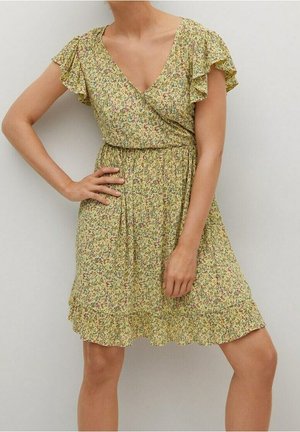 Femme portant une robe jaune à fleurs mi-longue avec des manches courtes volantées et un col en V, posant avec une main sur la hanche.