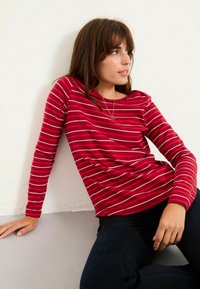 Red long-sleeve top with white horizontal stripes, round neck, and fitted design. Worn with dark jeans, against a textured white wall.