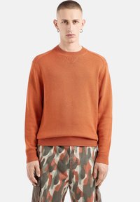 Oranger Strickpullover mit gerippten Bündchen und Saum, ausgestattet mit einem Rundhalsausschnitt und subtiler Textur, kombiniert mit Hosen im Tarnmuster.