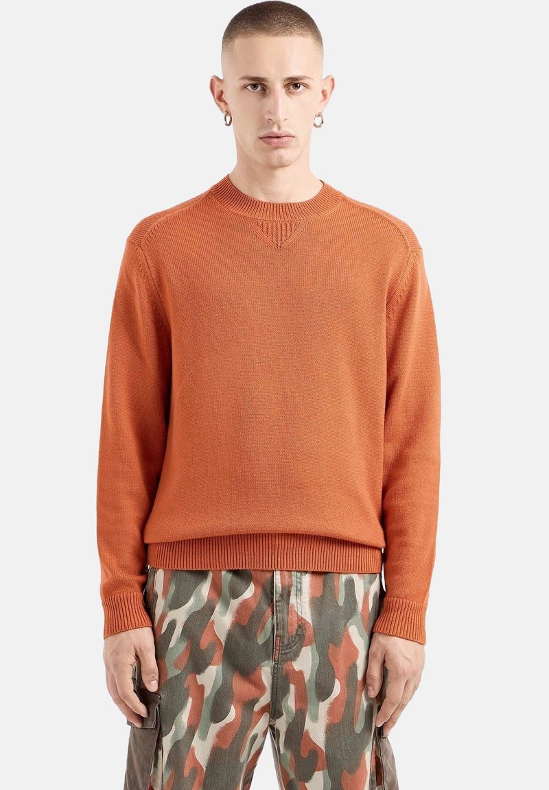 Oranger Strickpullover mit gerippten Bündchen und Saum, ausgestattet mit einem Rundhalsausschnitt und subtiler Textur, kombiniert mit Hosen im Tarnmuster.