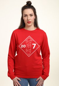 Červená mikina s dlouhými rukávy, s bílým grafick�ým designem skateboardu a textem "20 7" uvnitř diamantového tvaru. Materiál z bavlněné směsi.
