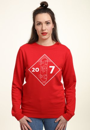 Czerwona bluza z długim rękawem, z białym wzorem graficznym przedstawiającym deskorolkę oraz napisem "20 7" w kształcie diamentu. Materiał mieszanki bawełny.