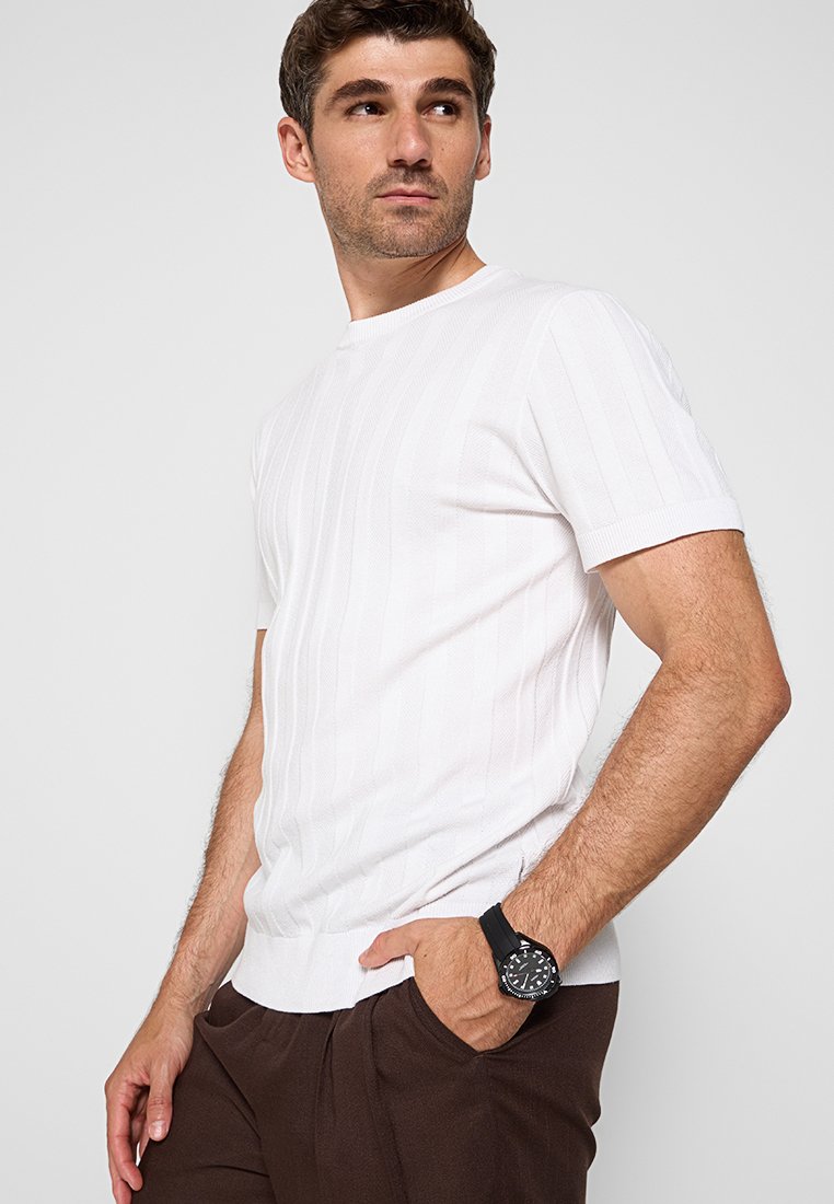 Weißes kurzärmliges Rippstrickshirt mit Rundhalsausschnitt und subtilen vertikalen Streifen, kombiniert mit dunkelbraunen Hosen und einer schwarzen Uhr.
