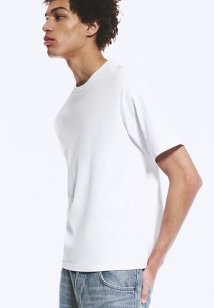 Tânăr cu păr creț purtând un tricou alb simplu și blugi albaștri, stând în profil cu mâna în buzunar.