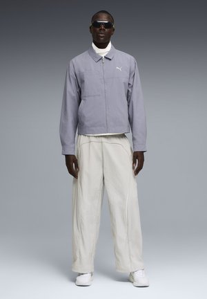 Man draagt een grijs jack met rits, een wit shirt met hoge hals, losse witte broek, witte sneakers en zwarte zonnebril en staat tegen een grijze achtergrond.