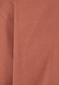 Țesătură de bumbac de culoare ruginie, cu o textură netedă, arătând cusături pe partea stângă, ceea ce indică un design de articol vestimentar casual.