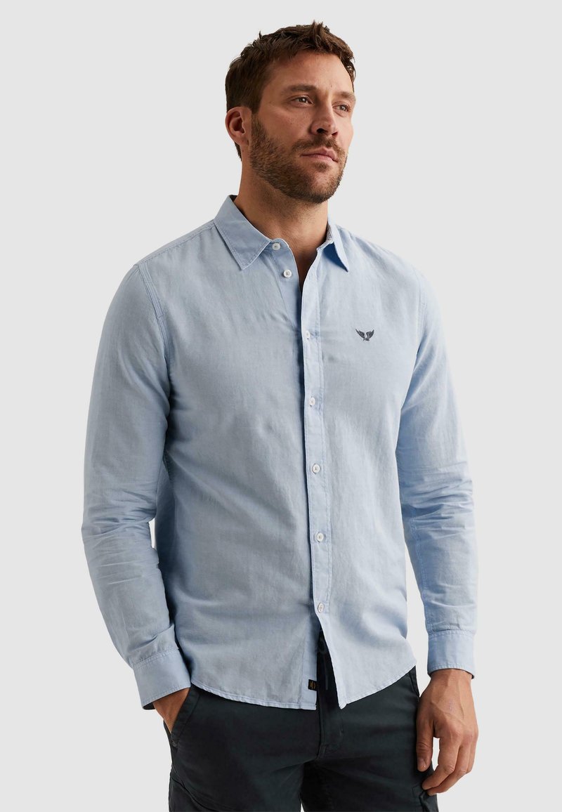 Man die een lichtblauw overhemd met knopen draagt en een klein geborduurd logo op de borst heeft, staand met zijn handen in de zakken van donkere broek tegen een effen achtergrond.