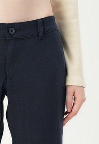 Pantaloni blu navy realizzati in tessuto testurizzato, con chiusura anteriore a bottone, passanti per cintura e tasche laterali. È visibile una manica di colore chiaro.