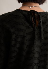 Schwarzes, strukturiertes Oberteil mit erhabenem, flauschigem Streifenmuster, das hinten mit einem Schlüssel-Loch-Verschluss aus Stoff gebunden ist und ein einzigartiges Designelement hinzufügt.